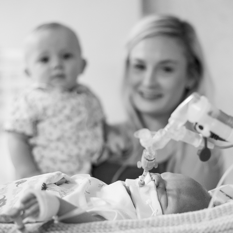 Out of focus mum and toddler gazing at a baby connected to a breathing tube in the foreground